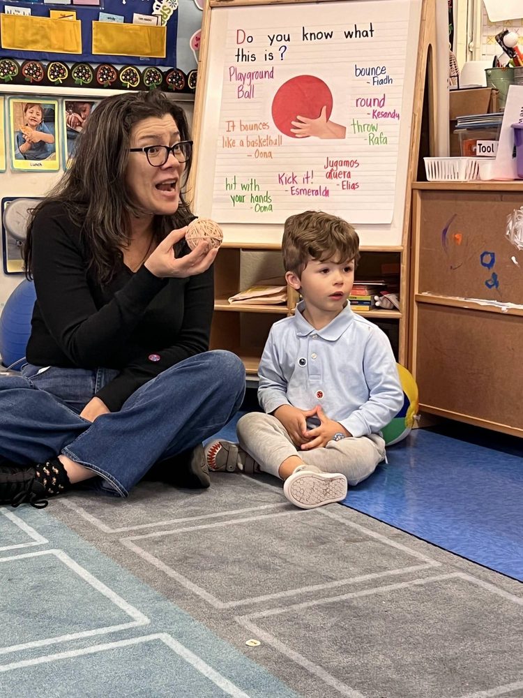 A woman demonstrates a ball game with a child seated beside her in a classroom.