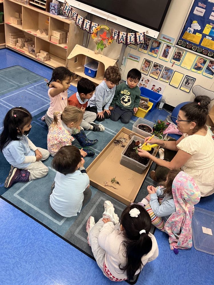 A teacher shows gardening activities to a group of children gathered around dirt trays.