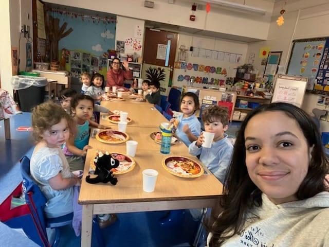 A group of children and adults enjoying pizza at a classroom table.
