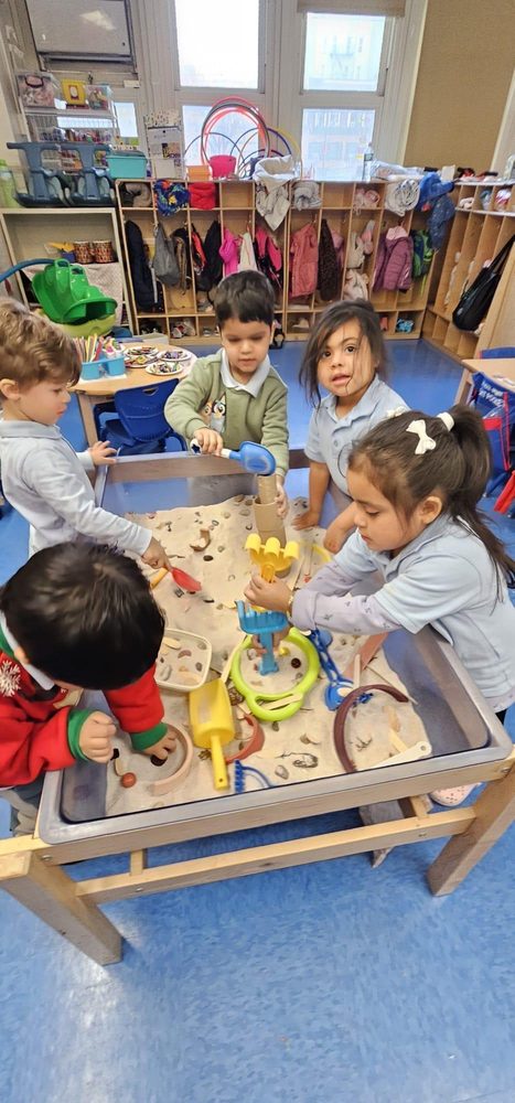 Two children play in a sand table, using toys with three other kids nearby in a classroom.