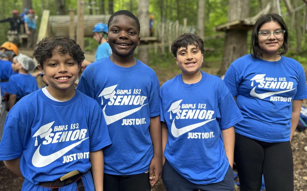 Four children smiling while wearing blue shirts in a green forest setting.