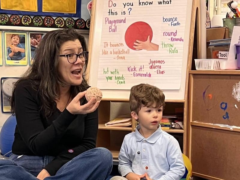 A teacher holds a small ball beside a young boy in a classroom.